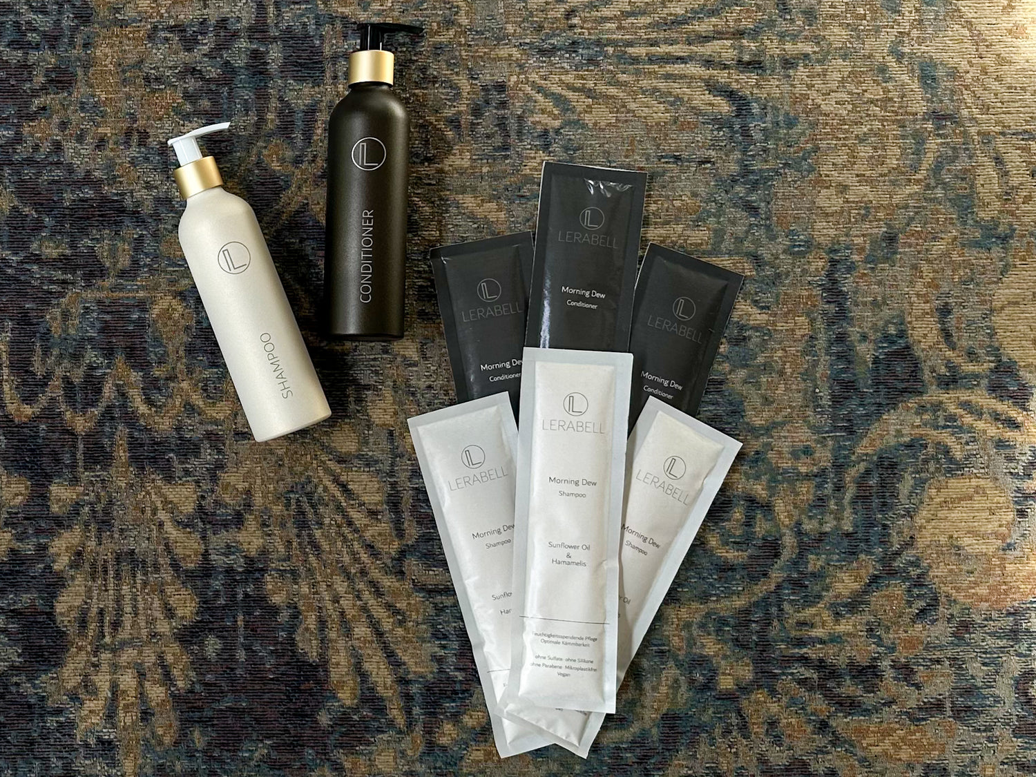 Lerabell Morning Dew Shampoo und Conditioner Stickpacks und Die beige und die braune Aluminiumflaschen mit Pumpaufsatz auf antikem Teppich von oben fotografiert
