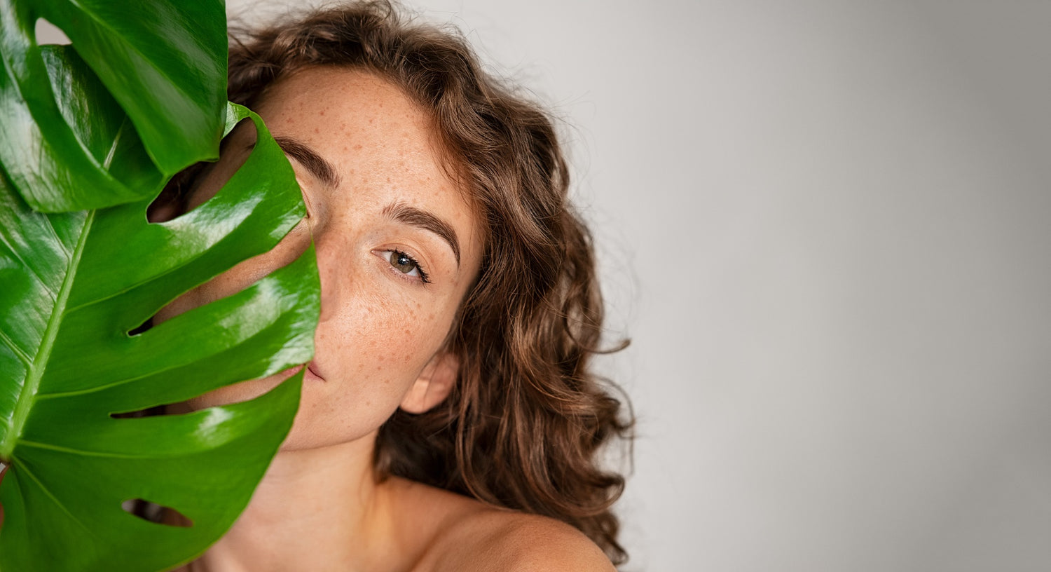 wunderschöne Frau mit Sommersprossen und braunen Haaren, versteckt ihr Gesicht zur Hälfte hinter dem Blatt einer Monstera
